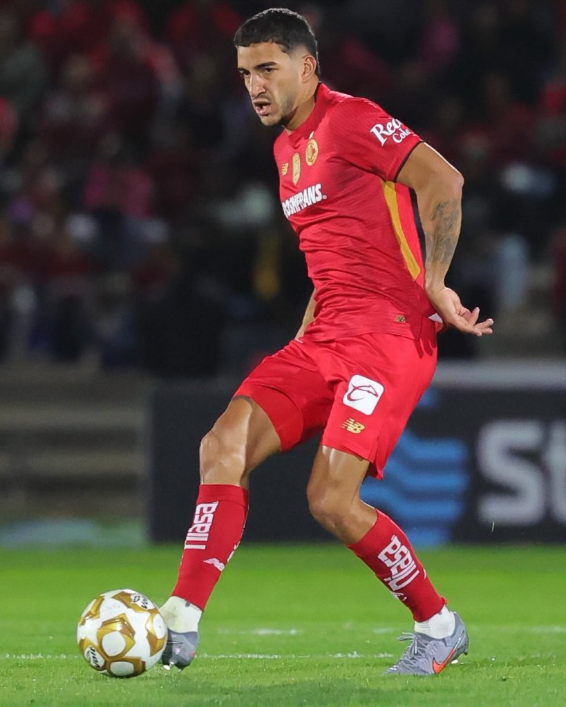 Un jugador de fútbol en uniforme rojo, realizando un pase con el balón en el campo.