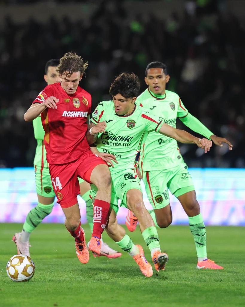 Un jugador con camiseta roja intenta driblar a un contrincante con camiseta verde durante un partido de fútbol, mientras otros jugadores se preparan para intervenir.