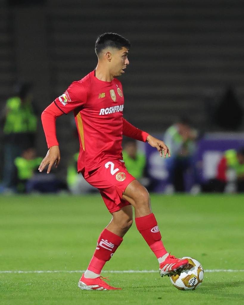 A soccer player in a red jersey and shorts dribbles a ball on the field, looking focused.