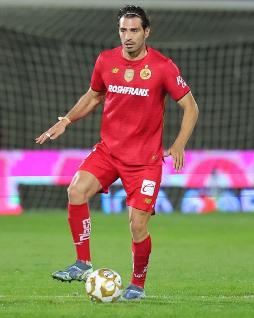 A soccer player in a red jersey stands on a field, preparing to kick a soccer ball. The player has short dark hair and is focused on the game.