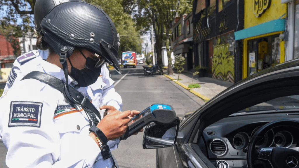 Una oficial de tránsito municipal con casco y máscara utiliza un dispositivo móvil para registrar una infracción vehicular en una calle de la ciudad.