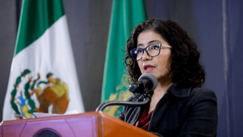Una mujer con cabello rizado y gafas habla en un atril rodeado de banderas. Un micrófono está frente a ella mientras presenta información relacionada con la Universidad Autónoma del Estado de México.