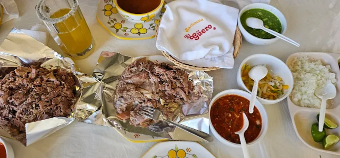 Mesa con platillos de barbacoa deshebrada, tortillas, salsas variadas y consomé caliente, acompañada de una bebida amarilla.