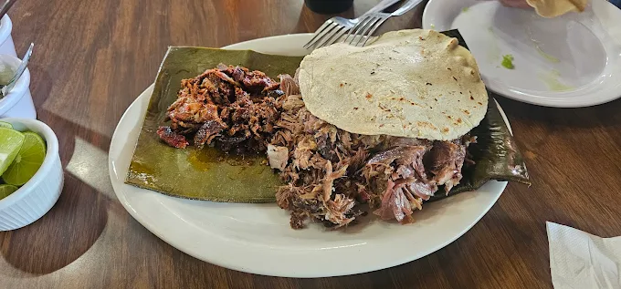 Plato de barbacoa servido con tortillas, carne deshebrada y salsa, acompañado de limones y cebollas en un restaurante.