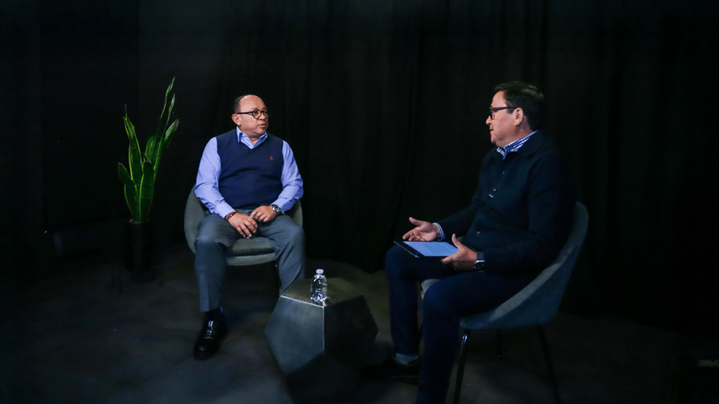 Dos hombres sentados en un estudio con fondo negro, uno de ellos lleva un chaleco azul y el otro una chaqueta oscura. Hay una planta verde decorativa y una botella de agua sobre una mesa entre ellos.