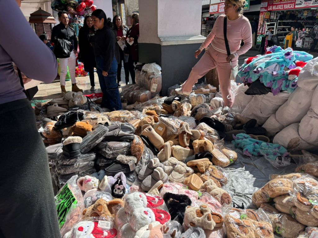 Mercado navideño en los Portales de Toluca, con productos como pantuflas y juguetes en el suelo, rodeado de compradores en un ambiente festivo.