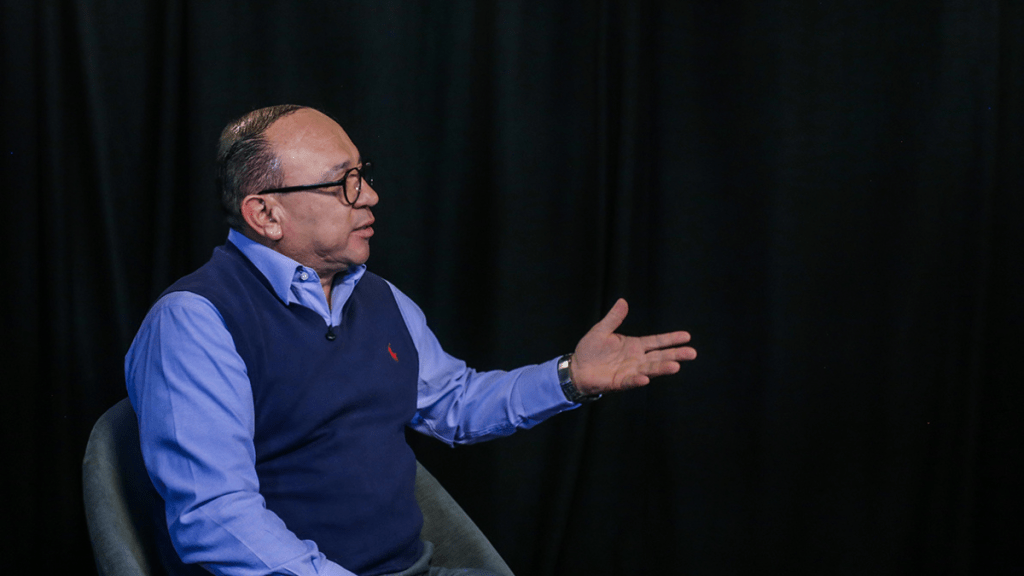 El alcalde de Zinacantepec, Manuel Vilchis Viveros, dialogando en un entorno interior, vestido con una camisa azul y un chaleco oscuro, gesticulando con una mano mientras habla.