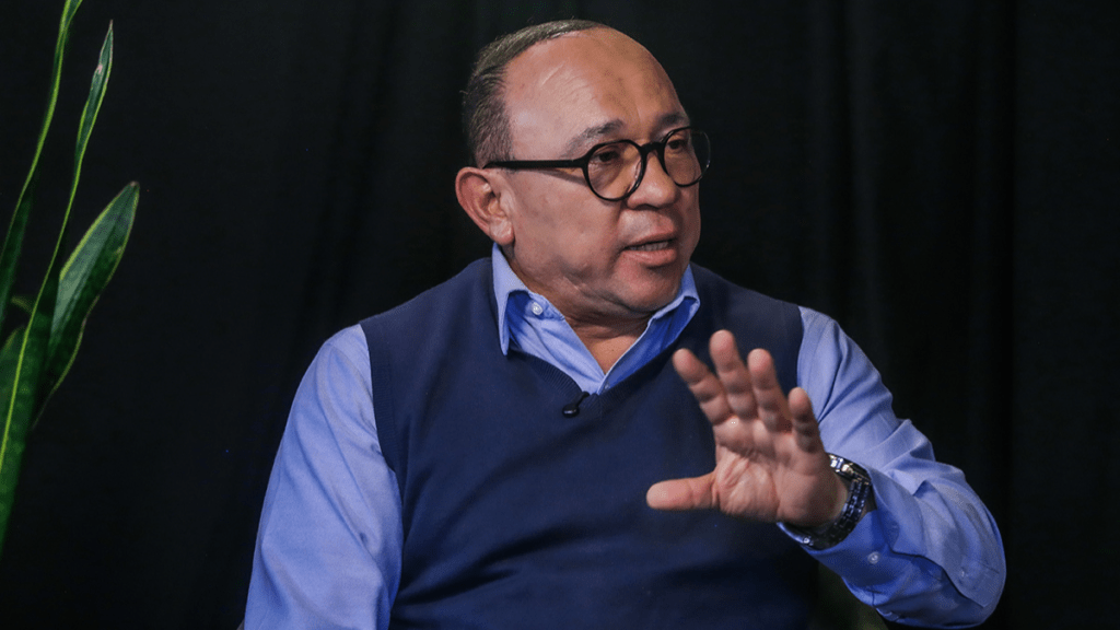 El alcalde de Zinacantepec, Manuel Vilchis Viveros, hablando en una entrevista, gesticulando con una mano mientras se sienta frente a una planta en un fondo oscuro.