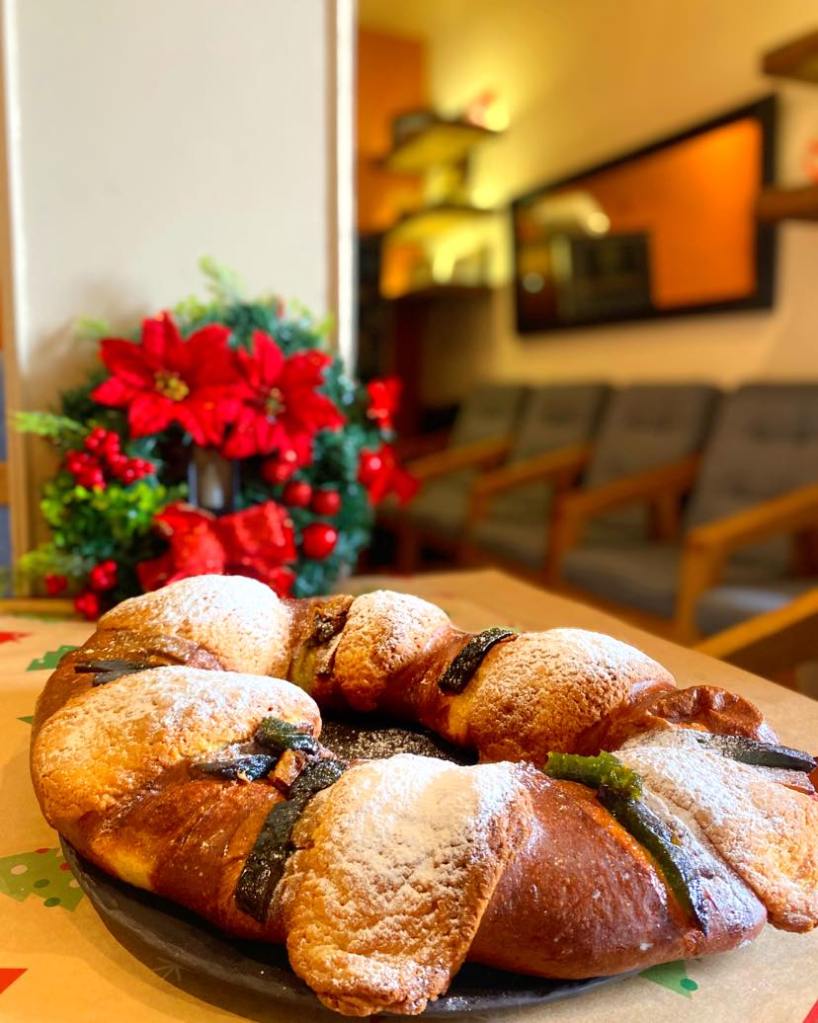 Rosca de Reyes decorada con azúcar en polvo y piezas de fruta, colocada sobre una mesa con un ambiente navideño de fondo.