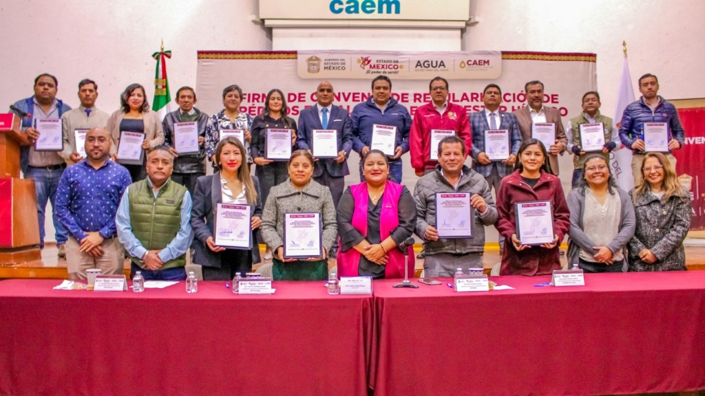 Grupo de personas posando con documentos en un evento de firma de un convenio de regularización fiscal en el Estado de México, con un fondo que incluye un cartel relacionado con el agua.