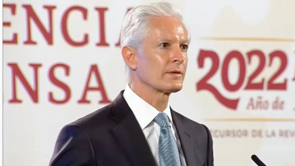 Retrato de un hombre con cabello blanco, vistiendo un traje oscuro y una camisa blanca, hablando en un evento formal ante un fondo con textos en rojo y dorado que incluye el año 2022.