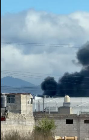 Nube de humo negro elevándose tras la caída de una avioneta en San Pedro Totoltepec, Toluca, con edificios y montañas al fondo.