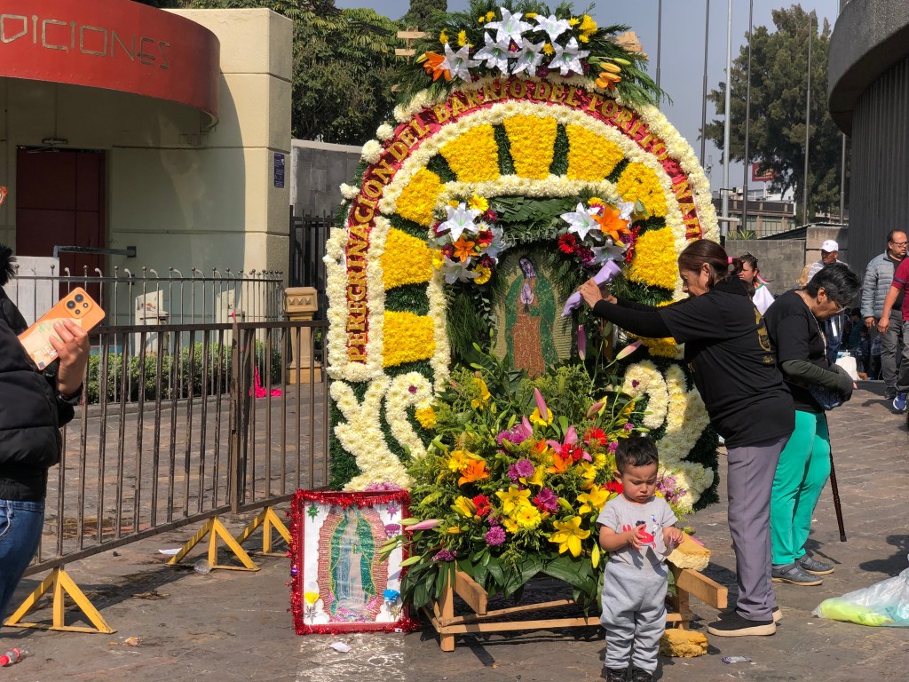 Grupo de personas decorando un altar lleno de flores en celebración del Día de la Virgen de Guadalupe, con un niño pequeño observando.