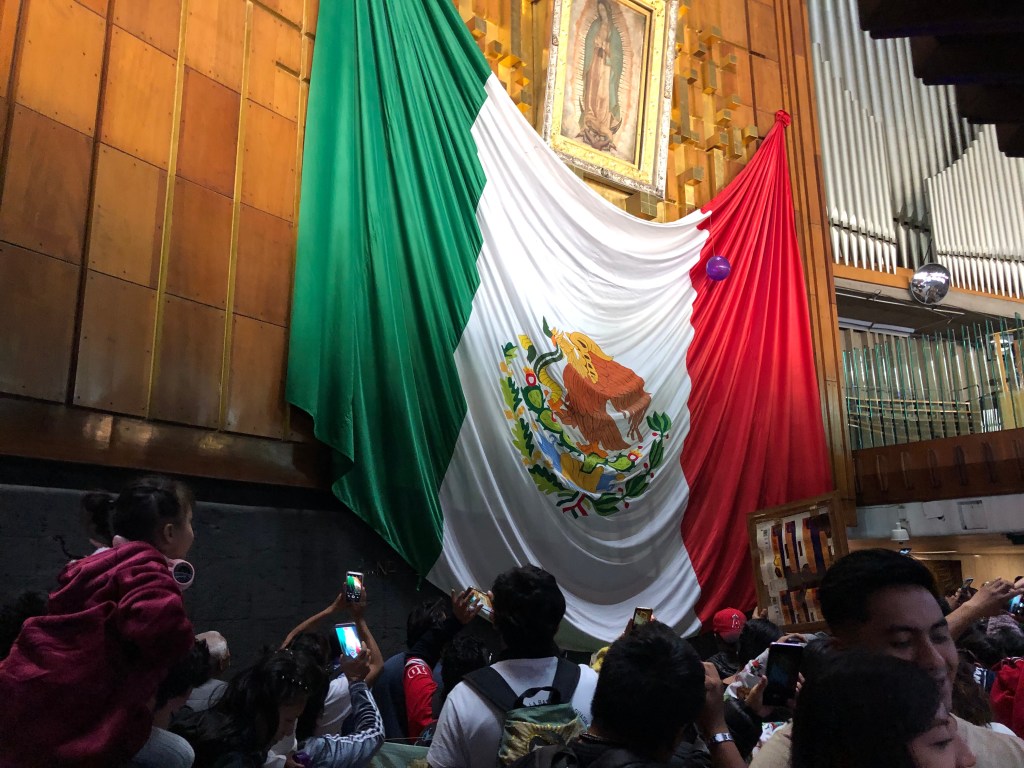 Interior de la Basílica de Guadalupe con un gran mural que representa la bandera de México, junto a la imagen de la Virgen de Guadalupe, mientras los fieles toman fotos y muestran su devoción.