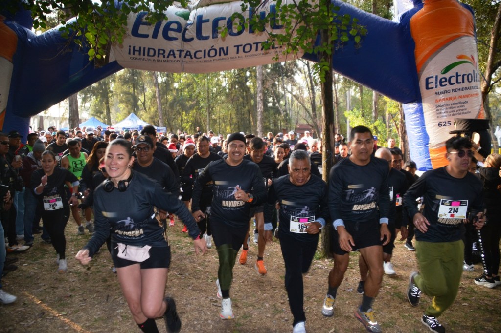 Participantes corriendo bajo un arco de inicio de carrera, vistiendo camisetas negras con el logo del evento en el Parque Naucalli durante la Primera Carrera Atlética Navideña 2025.