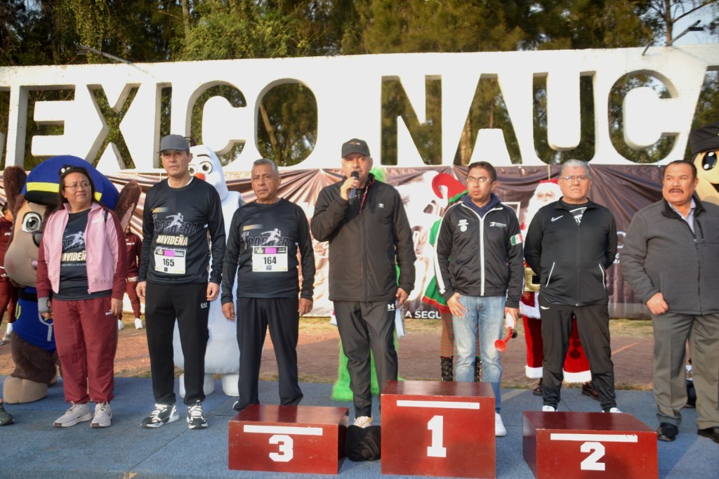 Grupo de personas en un evento deportivo, posando frente a una pancarta que dice 'MÉXICO NAUCALPAN'. Hay un hombre hablando al micrófono y varios participantes con camisetas de la carrera, junto a personajes de un disfraz, en un ambiente festivo.