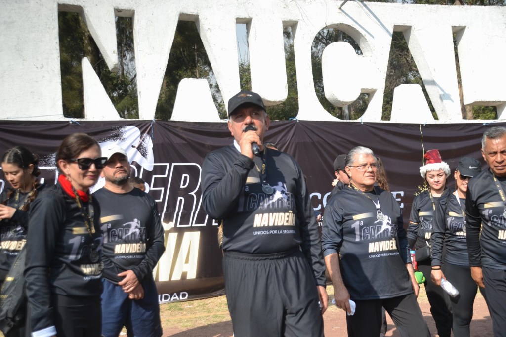 Un grupo de personas participa en la **Primera Carrera Atlética 'Navideña' 2025**, en el Parque Naucalli, con un ambiente de entusiasmo. Un hombre habla por un micrófono mientras otros oyen atentamente, todos vistiendo camisetas de la carrera.