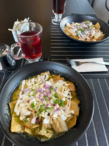 Plato de chilaquiles verdes servido en un bol negro, decorado con cebolla morada, crema y cilantro, acompañado de una bebida roja y otro plato de chilaquiles al fondo.