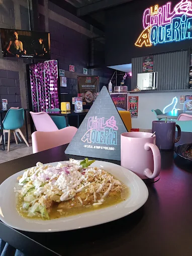 Plato de chilaquiles verdes decorado con lechuga y crema, servido en una mesa con decoración moderna y un letrero iluminado de fondo que dice 'La Chillaquería'.
