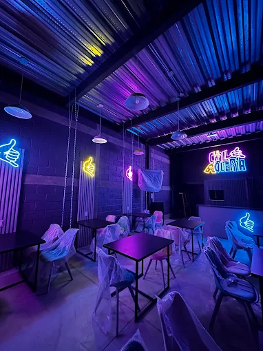 Interior de un restaurante con mesas y sillas cubiertas, decorado con luces de neón en colores vibrantes que incluyen un letrero que dice 'Chilaquería'.
