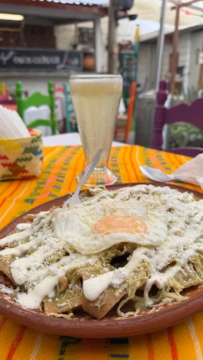 Plato de chilaquiles verdes con huevo estrellado, servido en un plato de barro, acompañado de una bebida en un vaso. Fondo colorido con manteles típicos mexicanos.