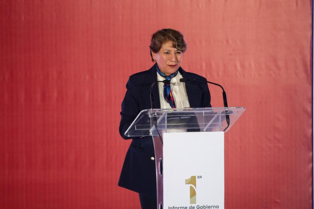 Una mujer de pie detrás de un podio en un evento formal, con fondo rojo y un cartel que dice '1er Informe de Gobierno'. Ella lleva un abrigo oscuro y una bufanda colorida mientras habla al público.