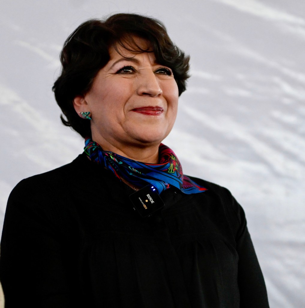 Delfina Gómez sonriendo mientras lleva un micrófono en un evento público, destacando su papel como gobernadora.