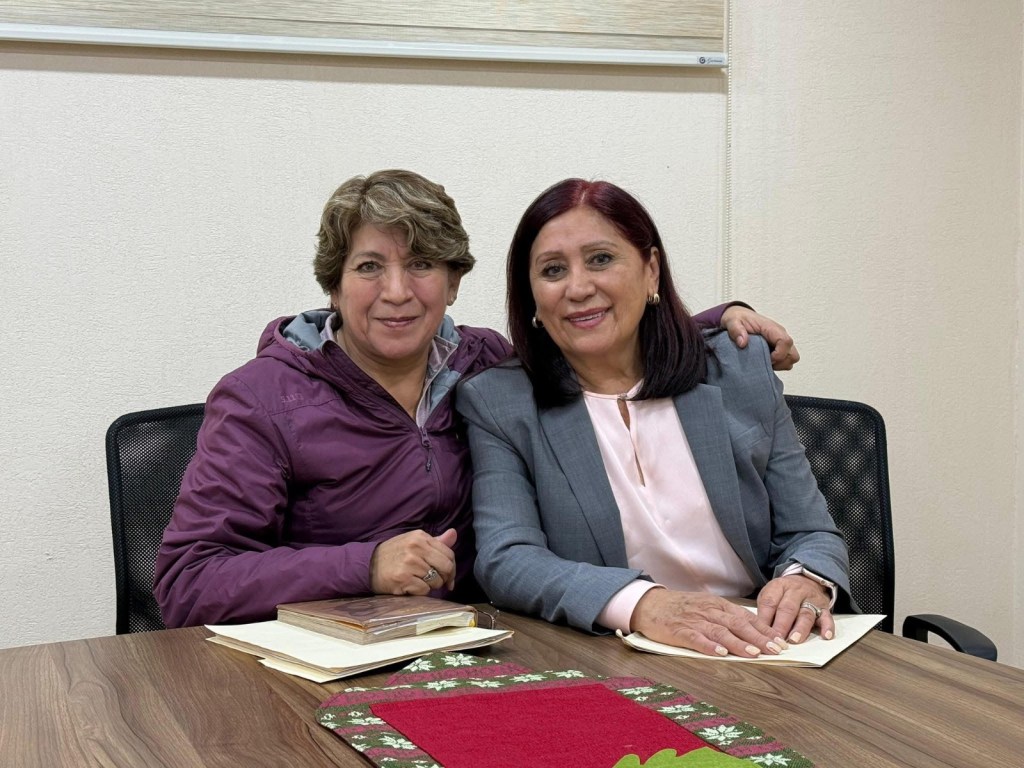 Dos mujeres sonrientes posando juntas en una mesa, con documentos y una decoración navideña visible.