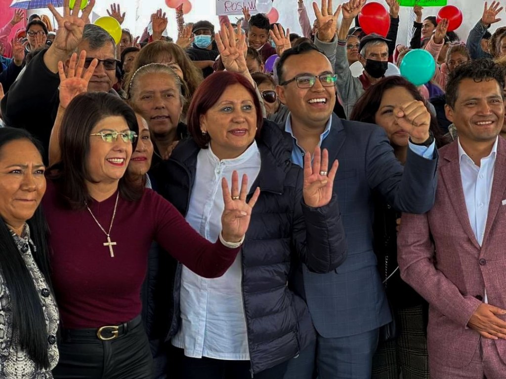 Grupo de personas sonrientes en un evento, levantando las manos con tres dedos extendidos, mostrando apoyo. Se puede ver un fondo con globos y carteles.