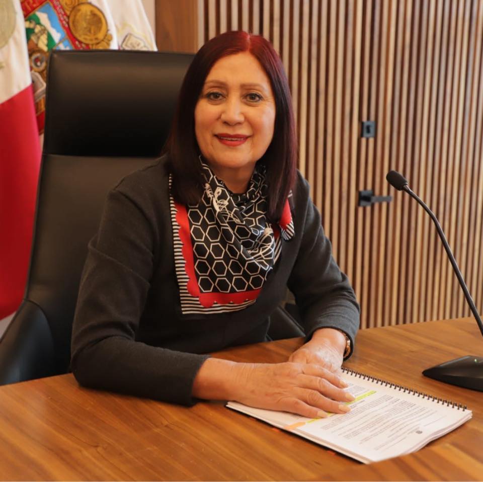 La alcaldesa de Tecámac, Rosa Yolanda Wong Romero, sonríe mientras está sentada en un escritorio con documentos y un micrófono a su lado.