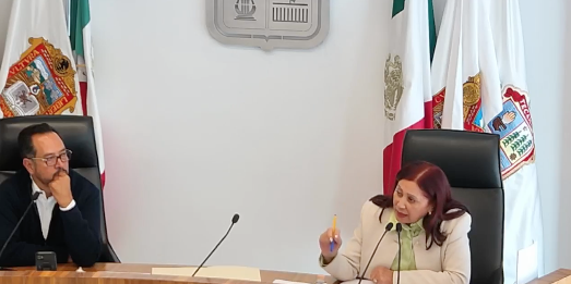 Reunión en el cabildo de Tecámac con la alcaldesa Rosa Yolanda Wong Romero y un colaborador, discutiendo temas administrativos. Se observan banderas y el escudo del municipio en el fondo.