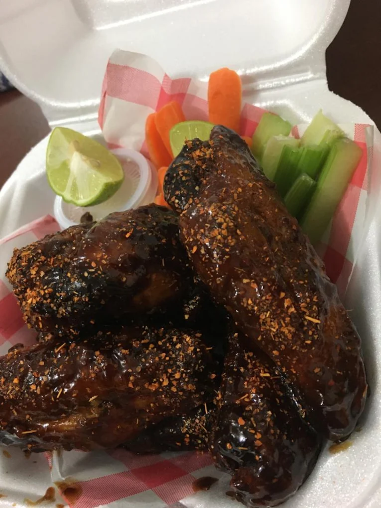 Alitas de pollo con salsa y especias, acompañadas de zanahorias, apio, rodajas de limón y un dip en un empaque de espuma.