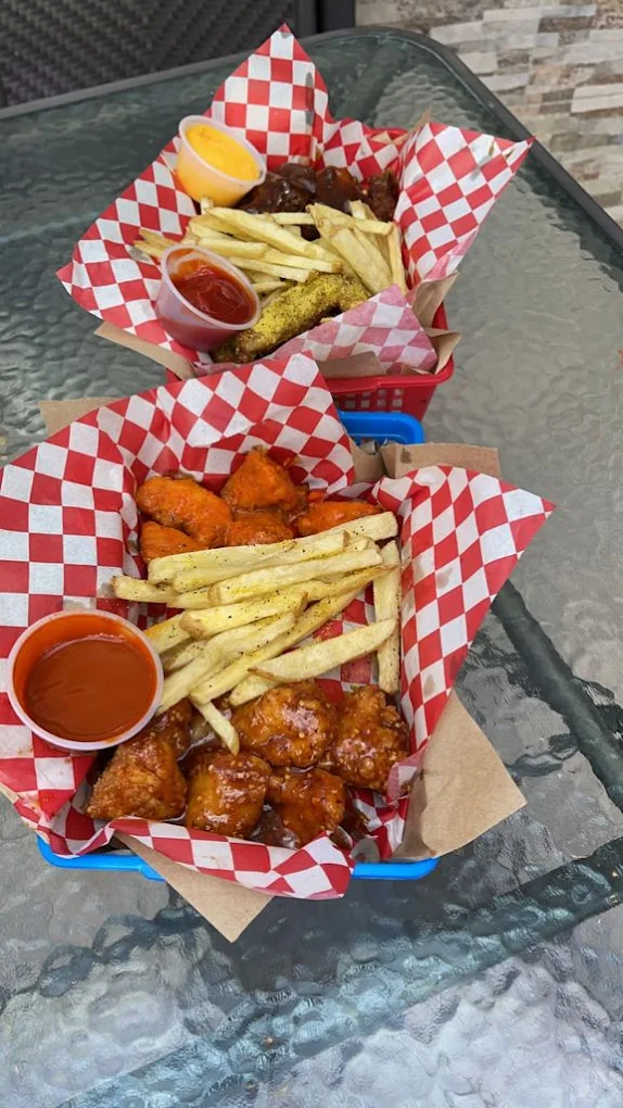 Platos de alitas con papas fritas y salsas, presentados en canastas con papel de cuadros rojos y blancos, sobre una mesa de vidrio.