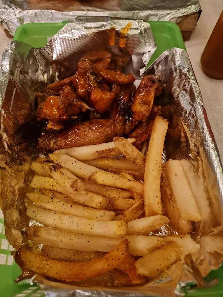 Caja con alitas de pollo sazonadas y papas fritas, servidas en un recipiente de papel aluminio.
