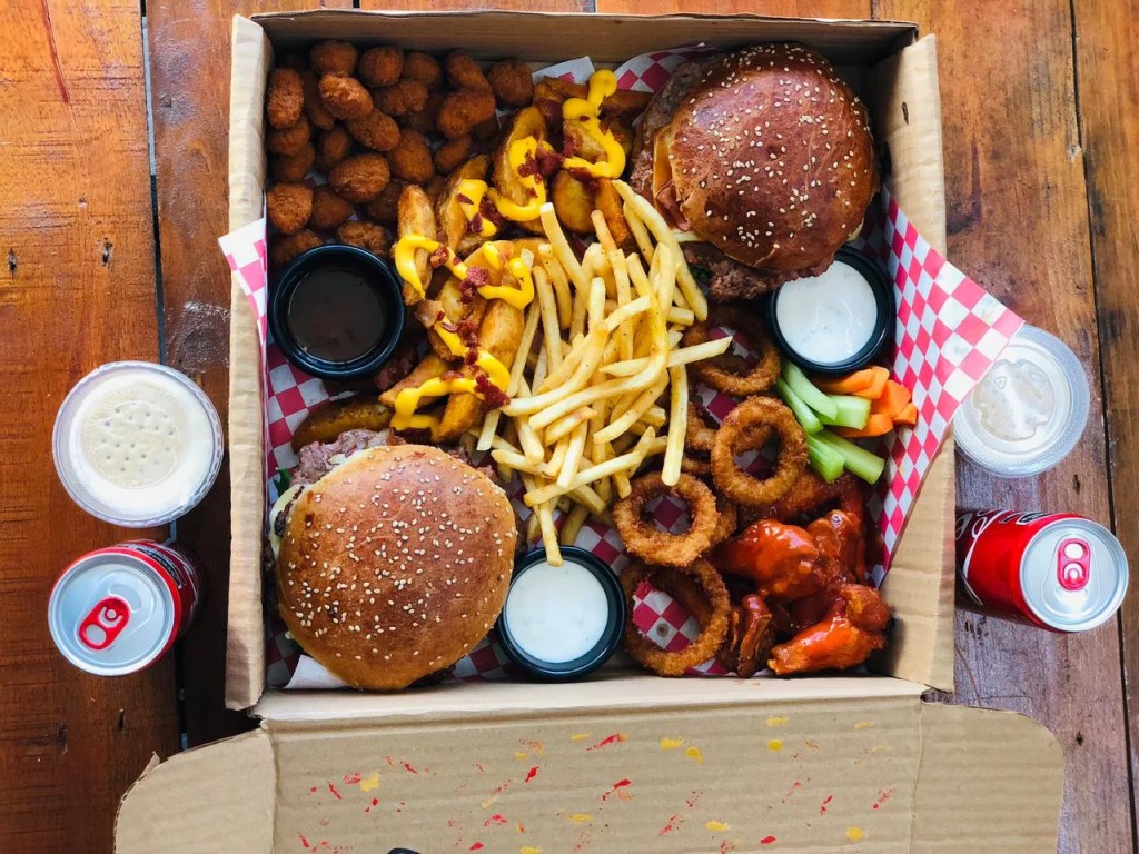 Un paquete de comida que incluye hamburguesas, alitas, papas fritas, aros de cebolla y una variedad de salsas en envase para llevar.