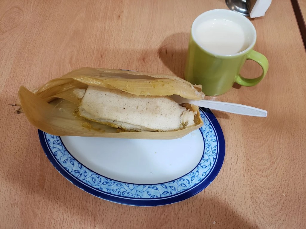 Tamale served in its corn husk on a plate, accompanied by a small cup of atole.