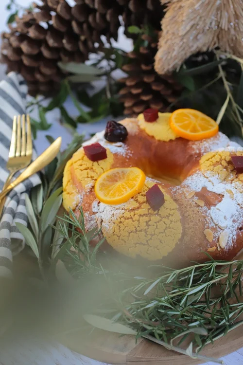 Rosca de Reyes decorada con rodajas de naranja, polvorón y otros adornos, sobre una base de hojas y piñas.
