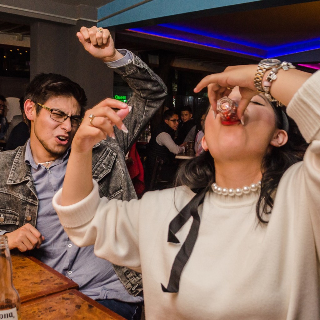 Dos personas en un bar celebrando con emoción, una de ellas levantando el brazo mientras la otra toma un trago, con un ambiente animado de fondo.