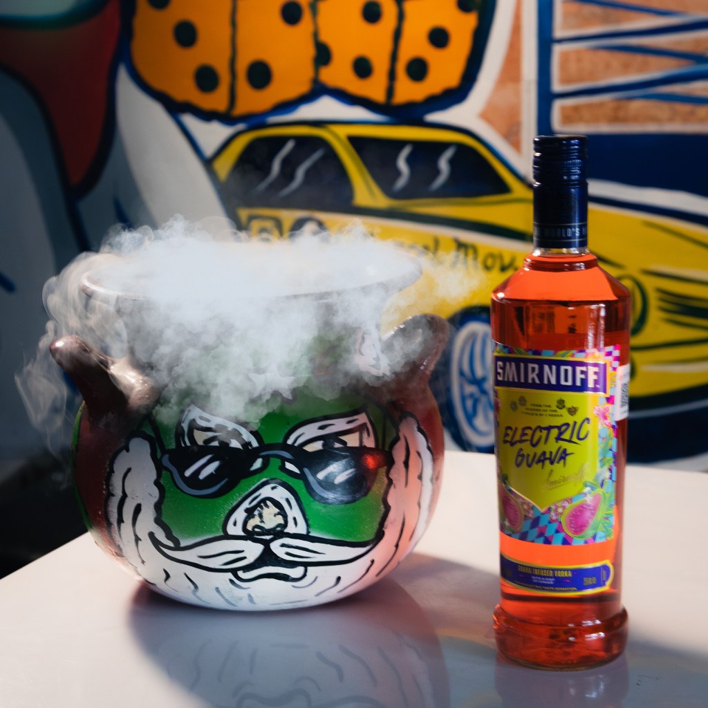 Un cóctel servido en una jarra con diseño de cara de Santa, emitiendo vapor, junto a una botella de vodka Smirnoff Electric Guava, con un fondo colorido.