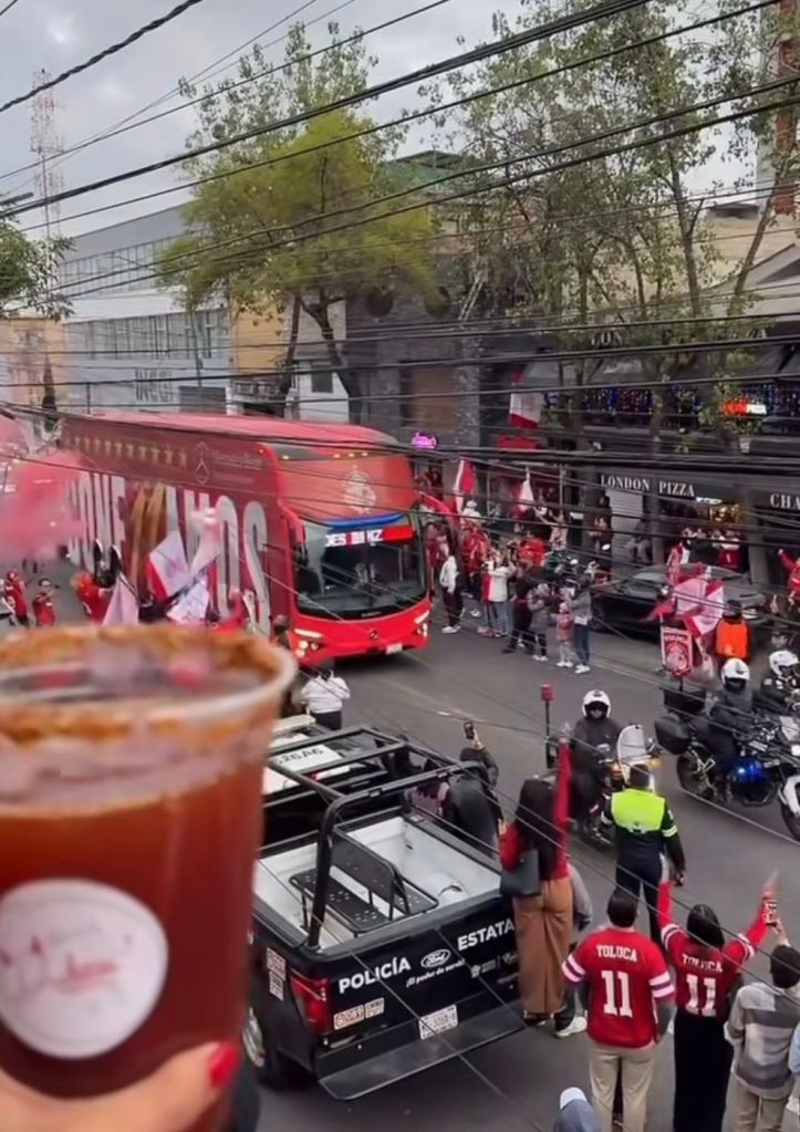 Una multitud de aficionados del Toluca con camisetas rojas celebrando en las calles mientras pasea un autobús de equipo, sosteniendo bebidas y animando con banderas.