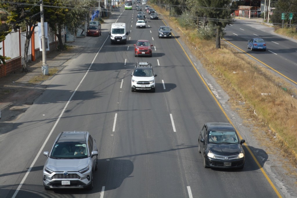 Vista de una carretera en el Estado de México, con varios vehículos en circulación. Se observa un camino dividido por líneas blancas y áreas de vegetación en los bordes.