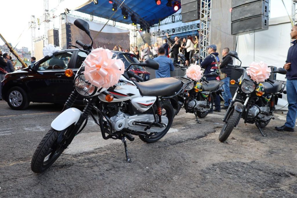 Motocicletas y un automóvil decorados con moños en un evento al aire libre, con una multitud de personas asistiendo al fondo.