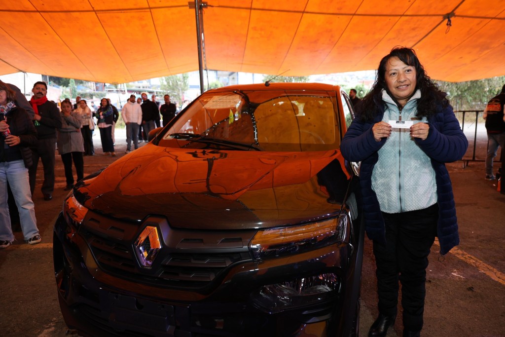 Una mujer sonriente sostiene un billete de sorteo frente a un automóvil negro, mientras una multitud se reúne detrás de ella bajo una carpa naranja.