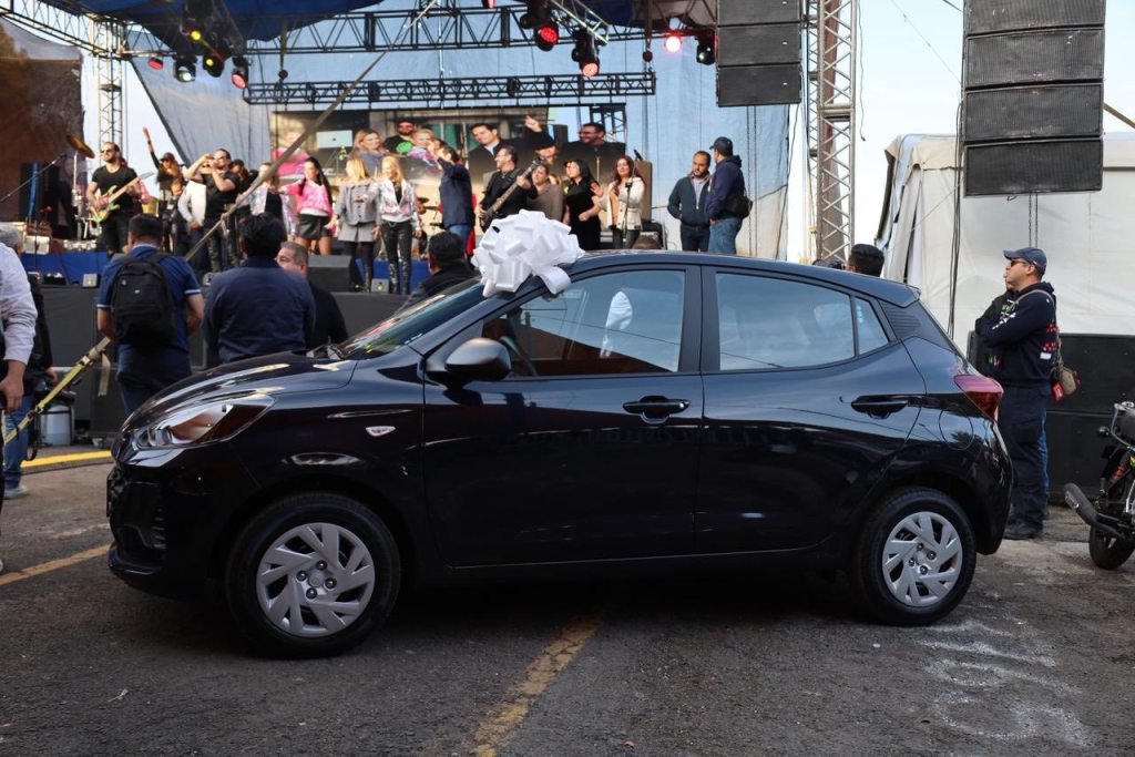 Un evento en Huixquilucan donde se presenta un automóvil negro con un lazo blanco, en un escenario con un grupo musical al fondo y una multitud de personas observando.