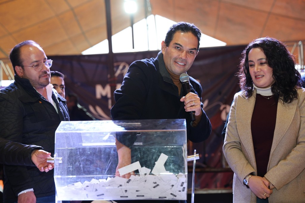 Tres personas participan en una rifa, una de ellas sostiene un micrófono mientras saca boletos de una urna transparente. Se observa un fondo con decoraciones para un evento.