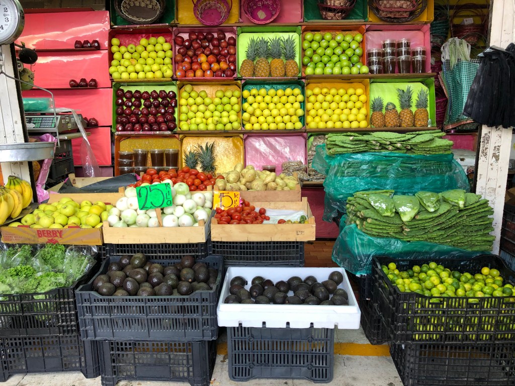 Un colorido puesto de mercado con frutas y verduras frescas, incluyendo plátanos, peras, tomates, aguacates y nopales, exhibidos en cajas con precios visibles.