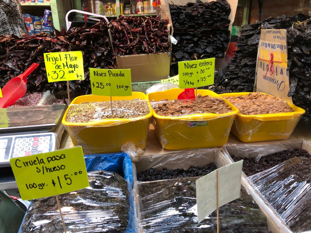 Vista de un mercado con varios recipientes amarillos que contienen productos como frijol flor de mayo, nuez picada, nuez entera y ciruela pasa, todos etiquetados con precios correspondientes.