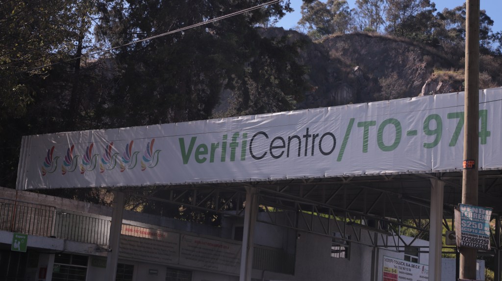 Exterior de un verificentro en Toluca con carteles visibles, incluyendo el nombre 'VerifiCentro/TO-974'. Se observa un entorno natural con árboles al fondo.