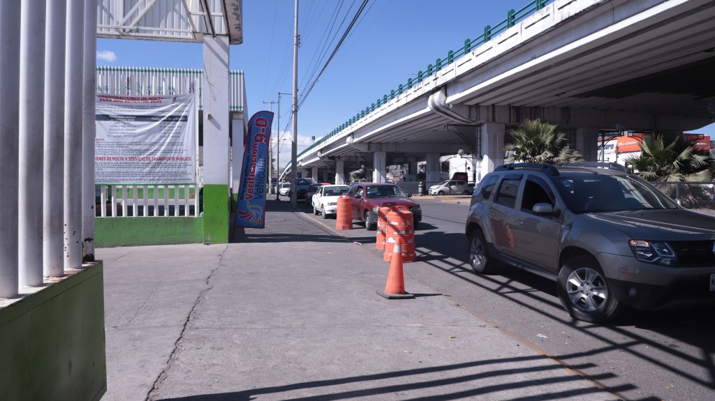 Vista de un verificentro en Toluca con vehículos en fila, señalización sobre la verificación y tráfico debajo de un puente elevado.