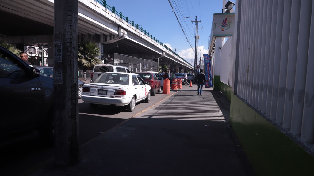 Vista de un verificentro en Toluca, con automóviles esperando en la fila y una persona caminando por la acera, bajo un puente.